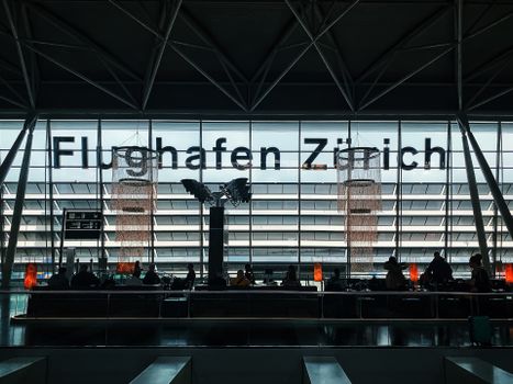 Travelers waiting at Zürich Airport with silhouettes against modern architecture.