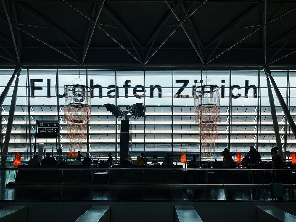 Travelers seated in silhouette against the tall windows of an airport waiting area