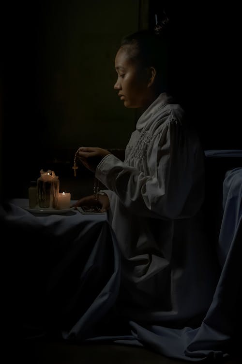 Free Side view of African American female teen in religious clothes with cross praying against flaming candles on black background Stock Photo