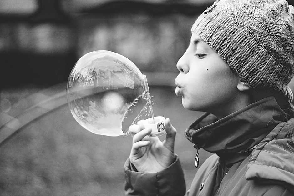 A child wearing a beanie blows a large soap bubble, captured in black and white.
