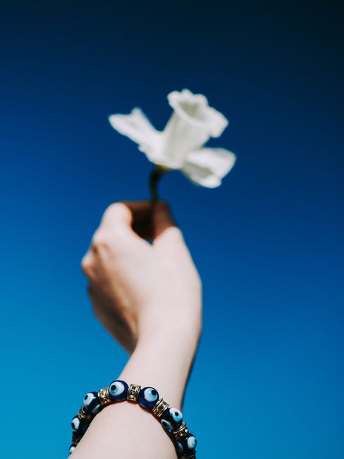 A hand holding a white flower with a bracelet against a vibrant blue sky.