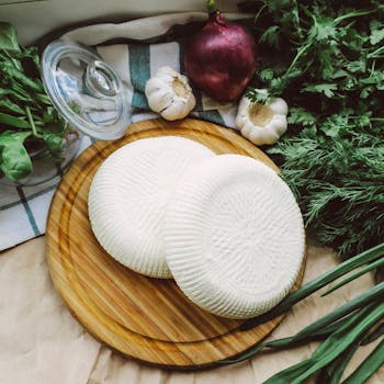 Top-down view of fresh cheese on a wooden board surrounded by herbs and vegetables.