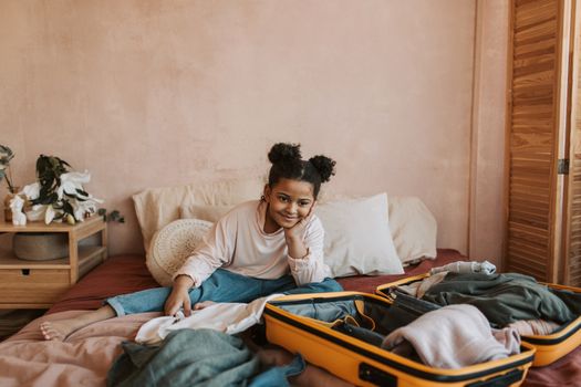 Young girl smiling while packing suitcase on a cozy bedroom bed.