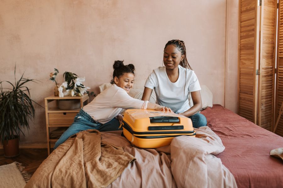 A mother and daughter share a joyful moment packing a suitcase on the bed.