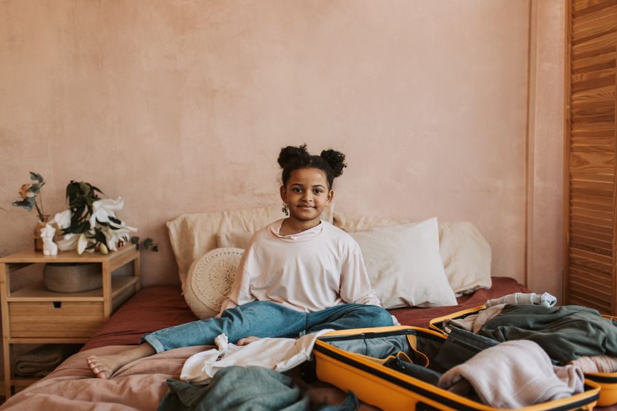 Smiling young girl sits on a bed while packing a suitcase in a warm, cozy bedroom.
