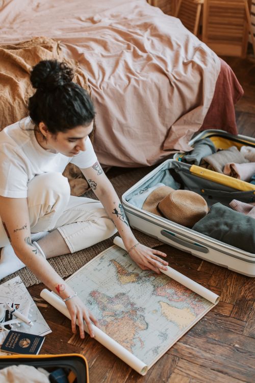 Free A young woman planning a travel adventure, seated by an open suitcase and unfolding a map. Stock Photo