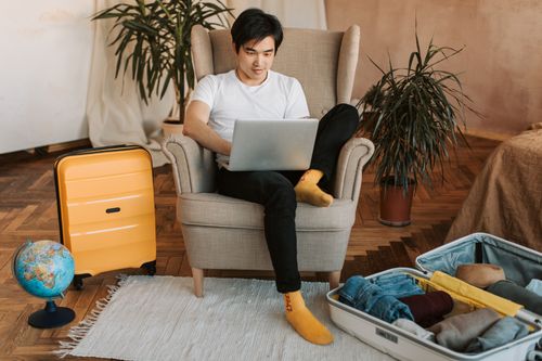 Free Man sitting on armchair planning travel, using laptop, with suitcases and globe nearby. Stock Photo