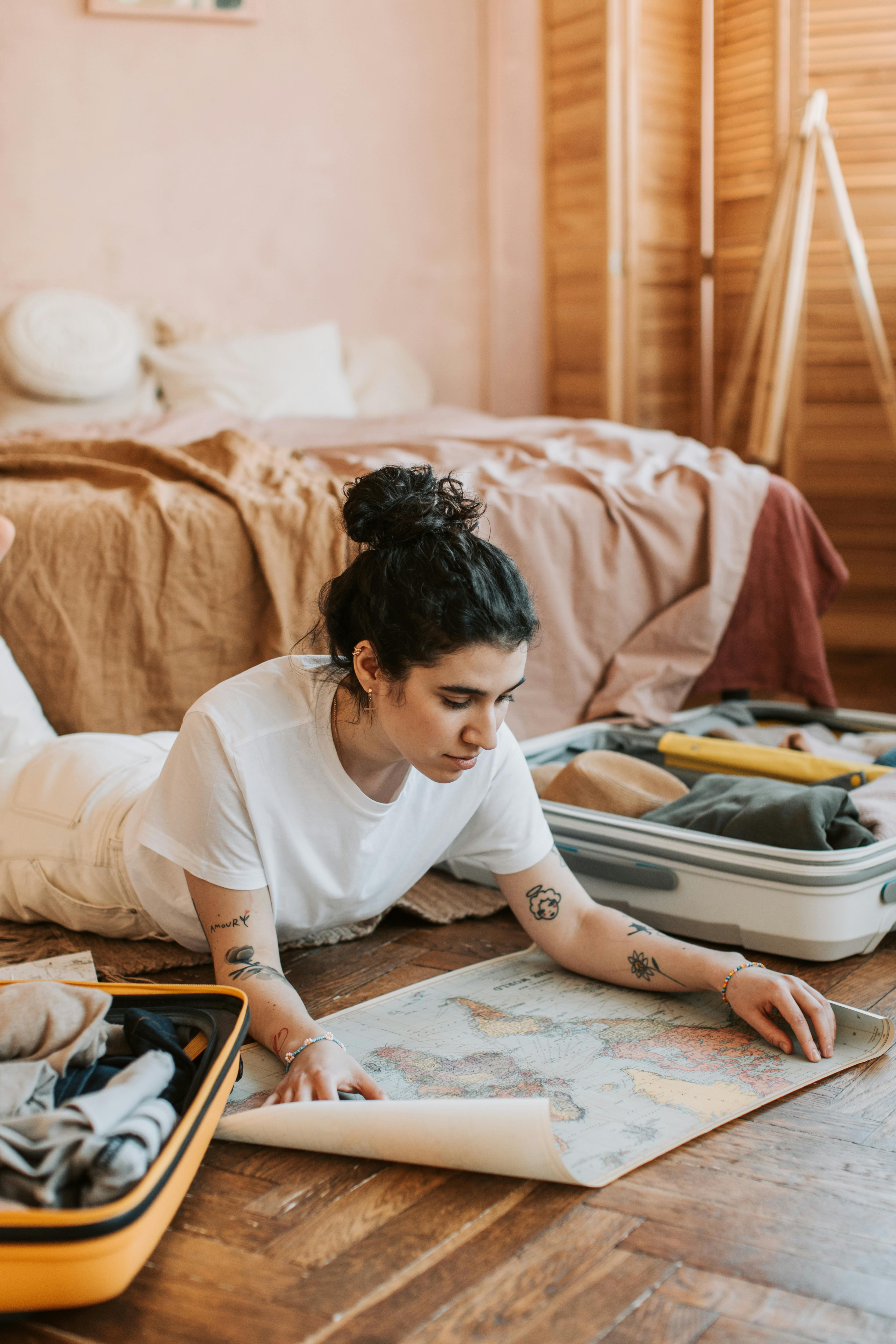 Tattooed woman in white shirt studies map near suitcase on wooden floor indoors. Travel preparation ambiance.
