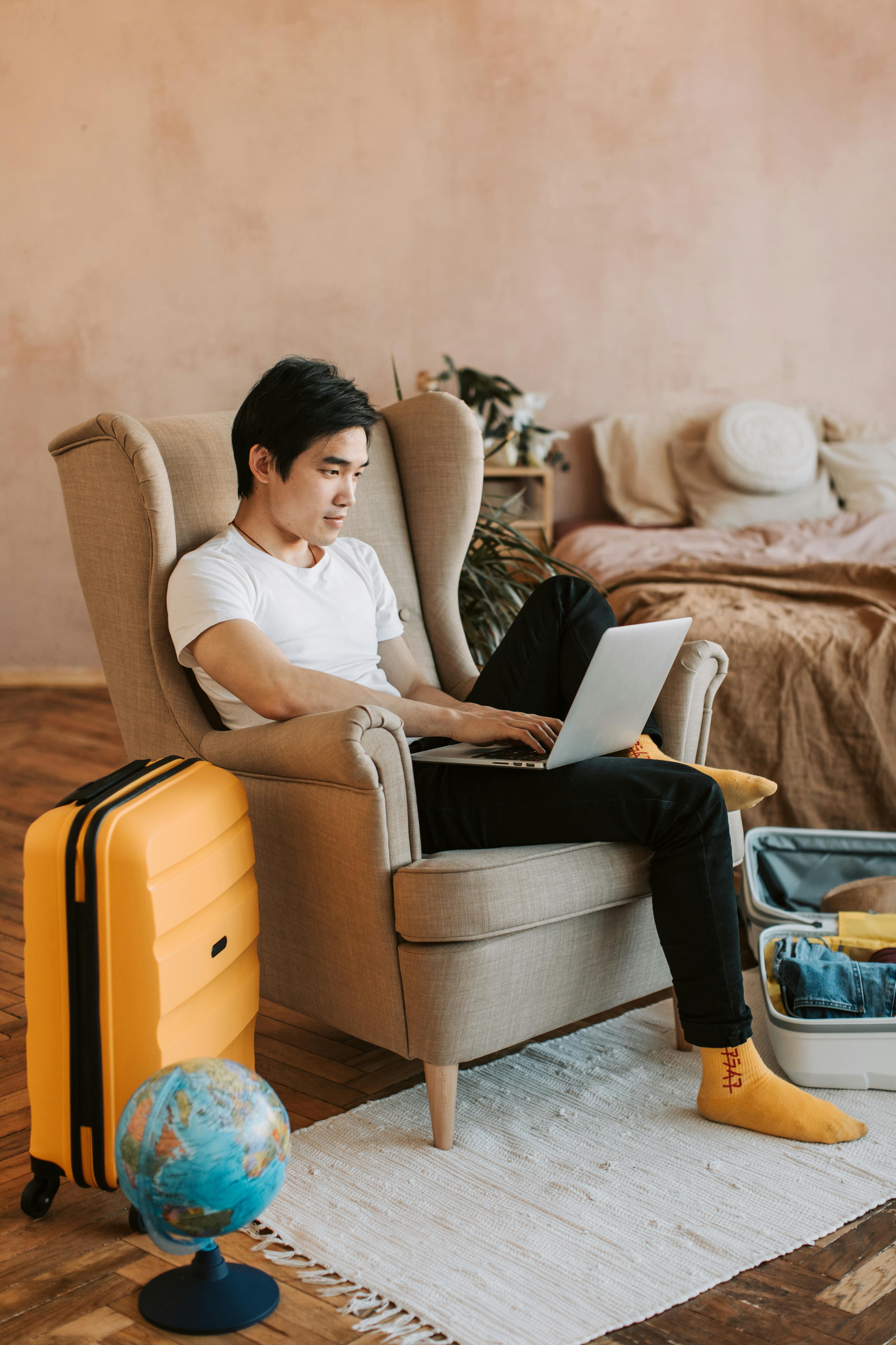 Asian man with suitcase and globe planning travel on a laptop at home.