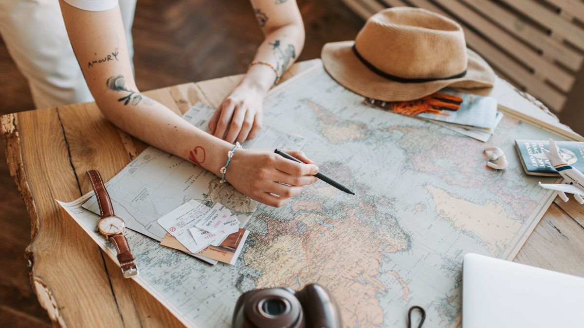Tattooed woman planning a trip with a world map, passports, and travel essentials on a wooden table.