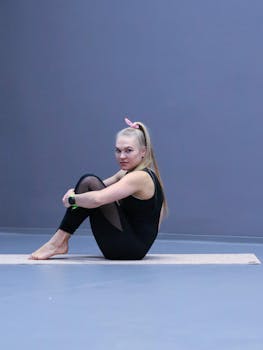 A young woman in fitness attire sitting on yoga mat indoors, looking at the camera.