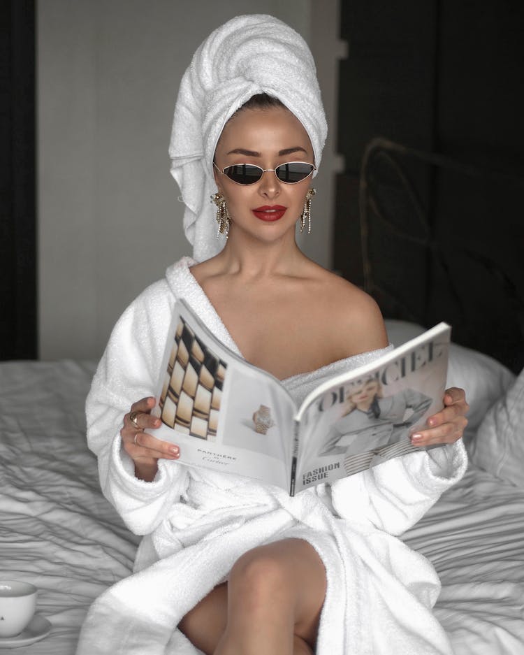 Woman Wearing A Bathrobe And With Hair Wrapped In A Towel Sitting In Sunglasses And Reading A Magazine 