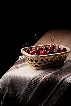 A wicker basket filled with vibrant red cherries on a wooden table with cloth, shot against a dark background.
