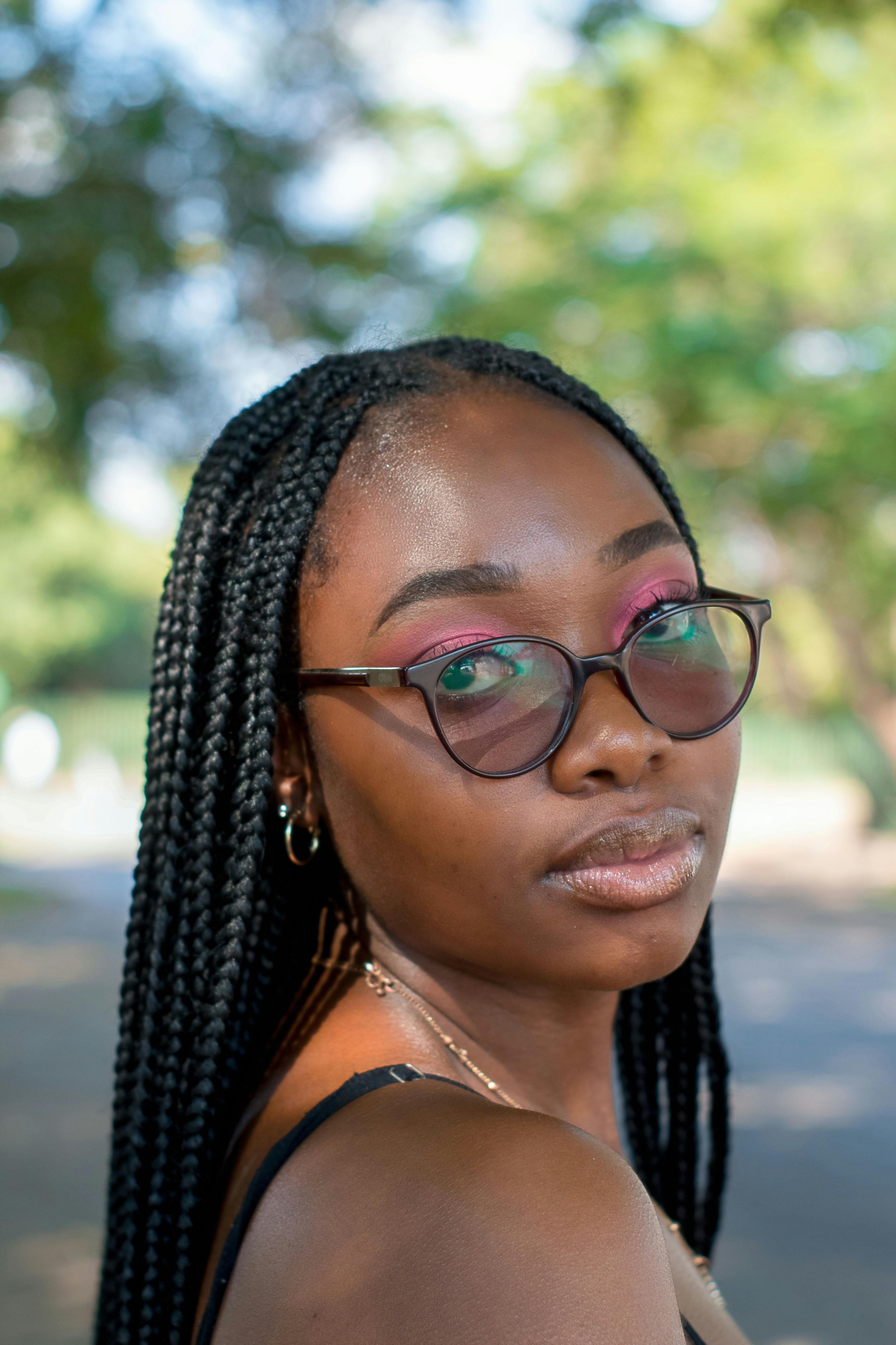 Close-up portrait of a woman with eyeglasses and braided hair enjoying sunlight outdoors.