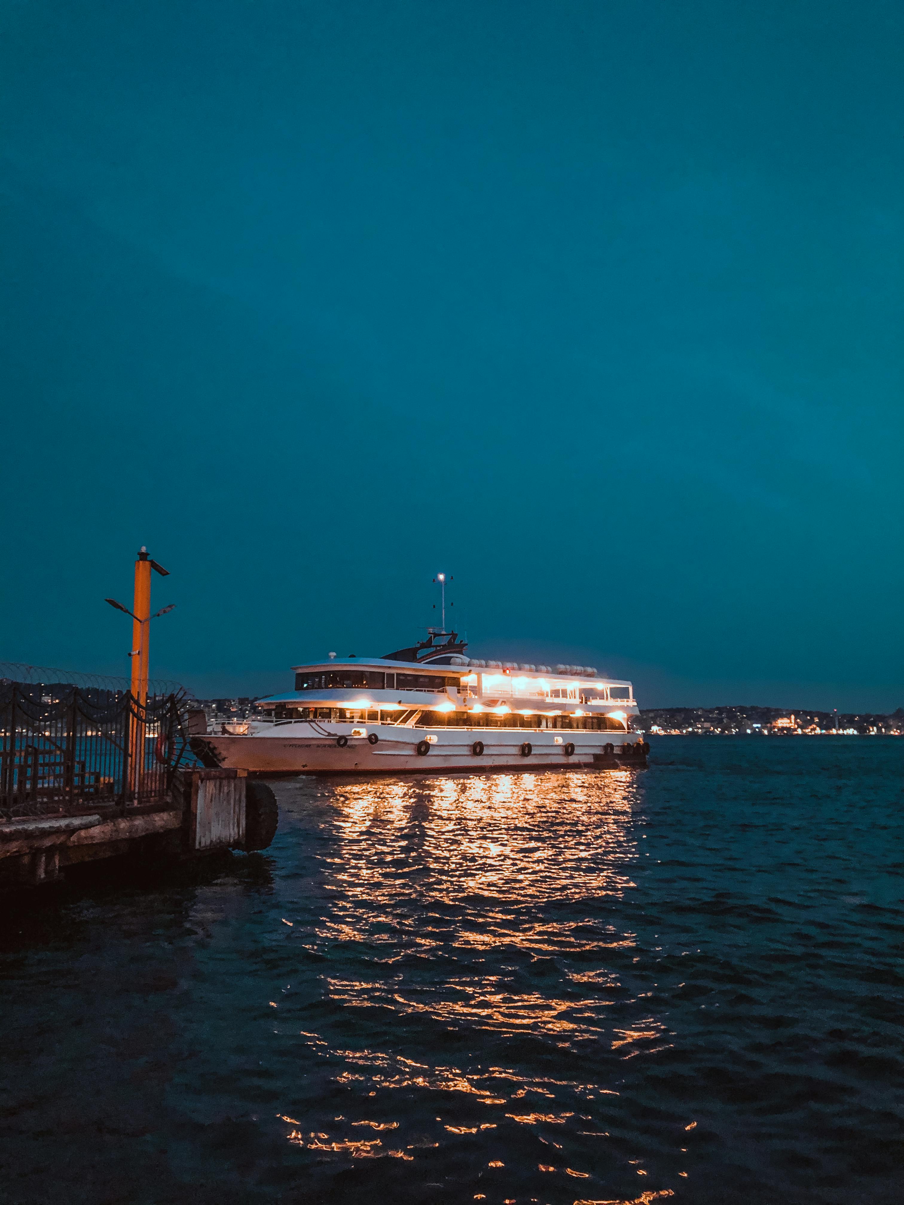 Free A beautifully illuminated ferry boat captured at dusk, reflecting off a calm sea with a city skyline in the background. Stock Photo
