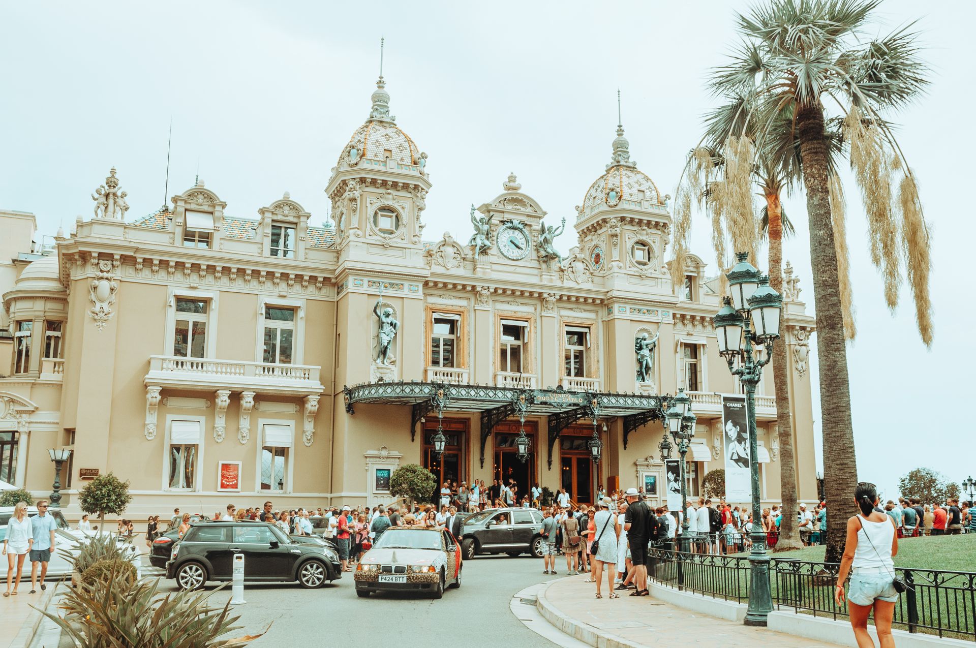 The iconic Monte Carlo Casino exudes grandeur with a bustling crowd and palm trees on a sunny day.