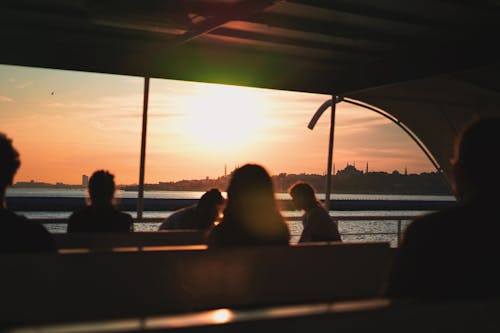 Unrecognizable people sitting on benches while sailing on sea against coastal city at sunset time during cruise trip on ferry