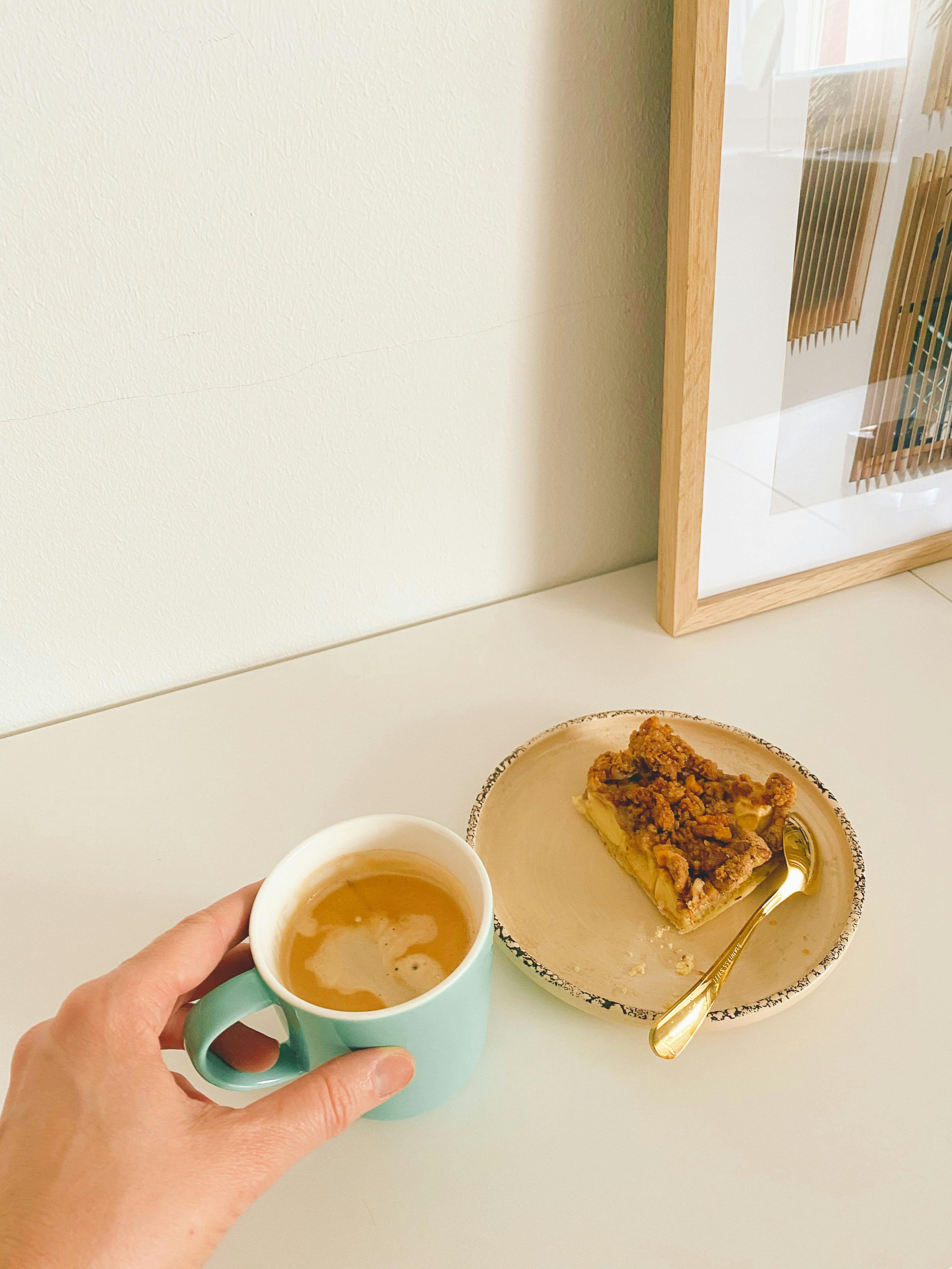 Female hand with ceramic mug of fresh aromatic cappuccino with piece of yummy homemade dessert placed on plate on table