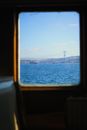Through window of modern vessel floating on rippling sea water against modern bridge near coastal town with residential buildings on summer day