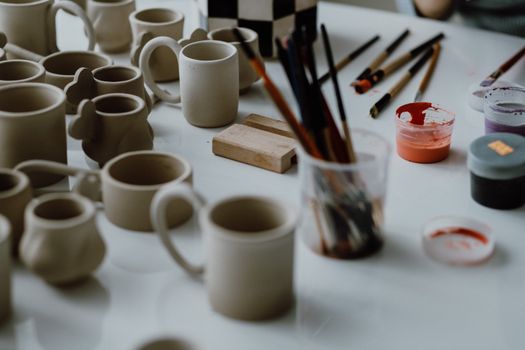 Artistic setup of pottery tools and unpainted ceramic mugs ready for glazing on a table.