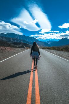 A woman walks alone on a mountain road under a vibrant blue sky, surrounded by scenic views.
