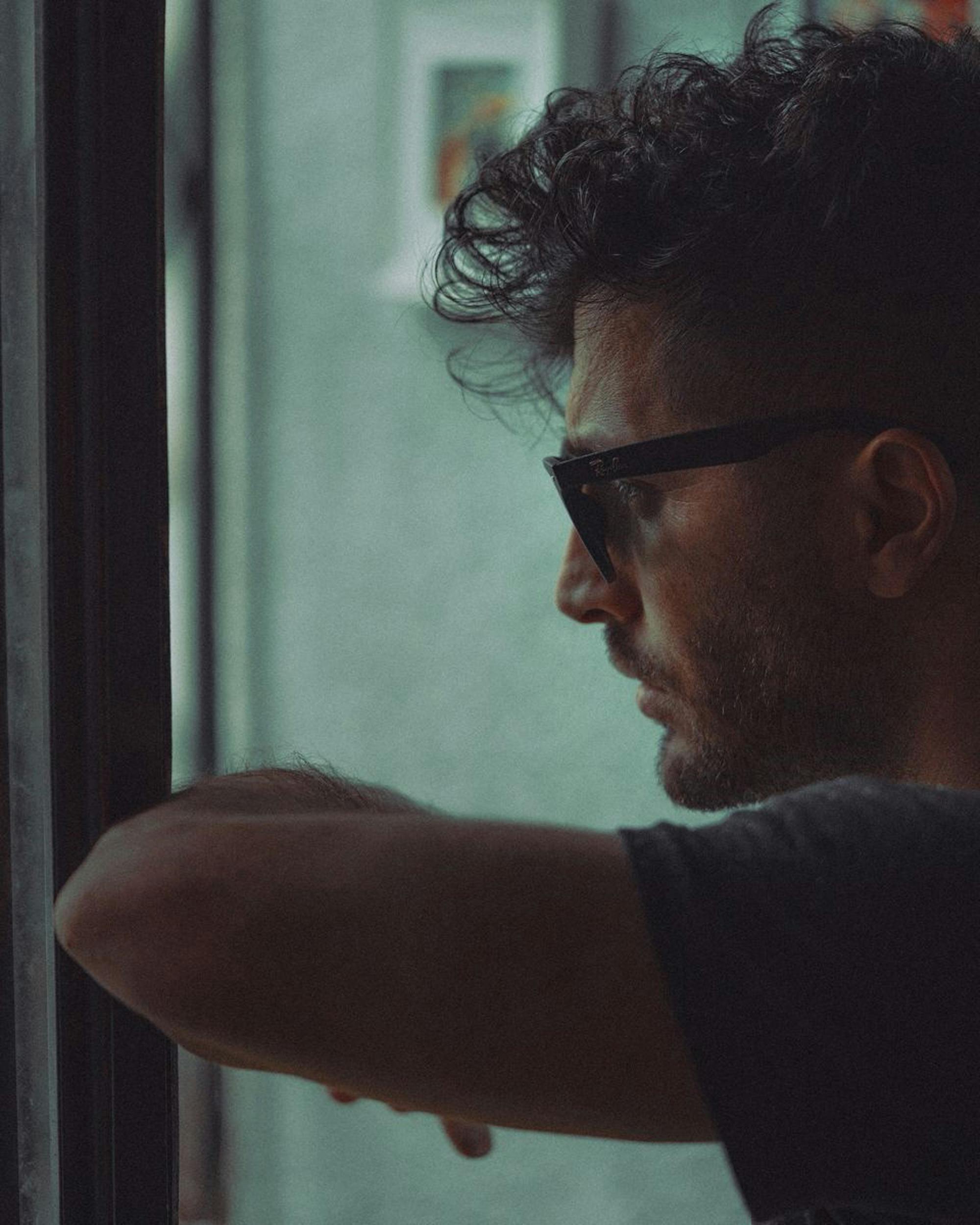 Close-up side profile of a man wearing sunglasses, thoughtfully gazing out a window.