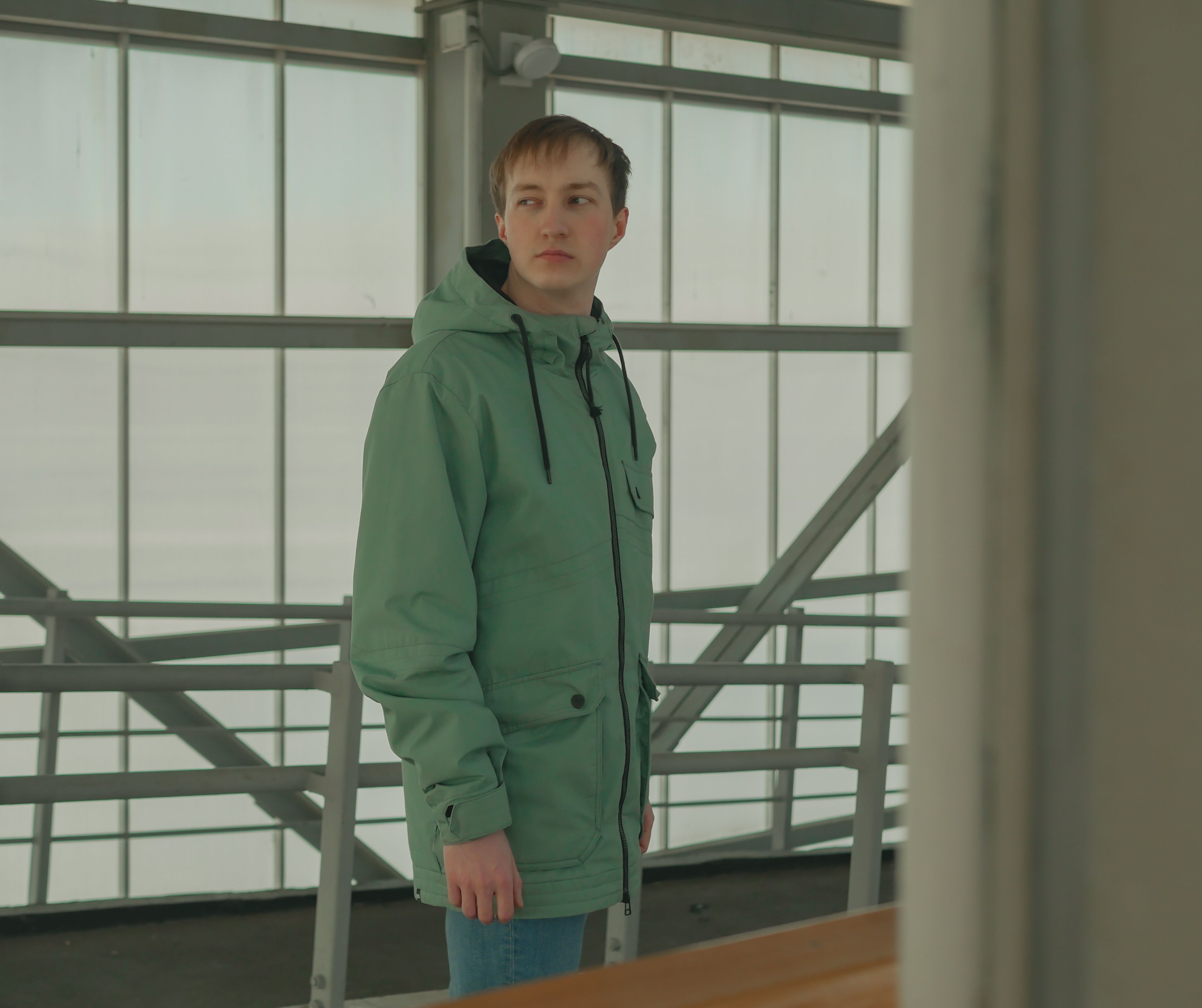 Man in green jacket posing indoors looking back with modern structure background.
