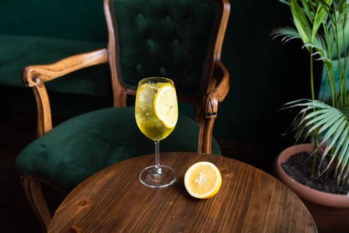 Free A refreshing lemon cocktail on a wooden table with a cozy, elegant chair and lush green plant. Stock Photo