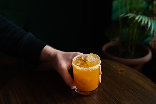 Free Close-up of a hand holding an orange cocktail with ice cubes on a wooden table indoors. Stock Photo