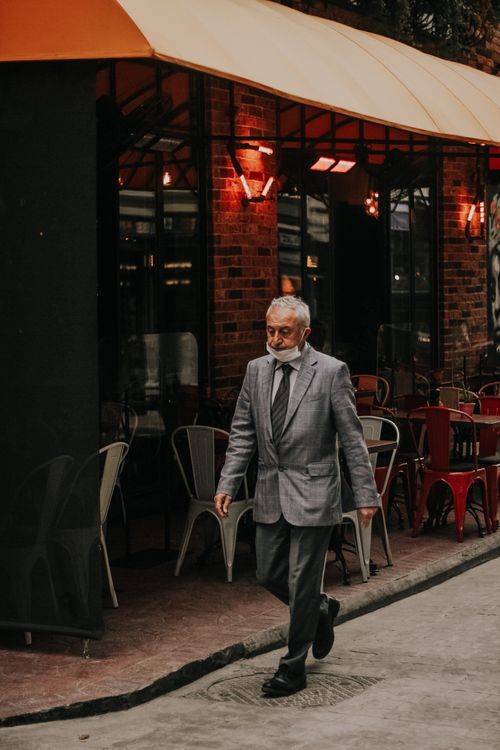 Free An elderly man in a gray suit walks past a stylish outdoor cafe with brick facade. Stock Photo