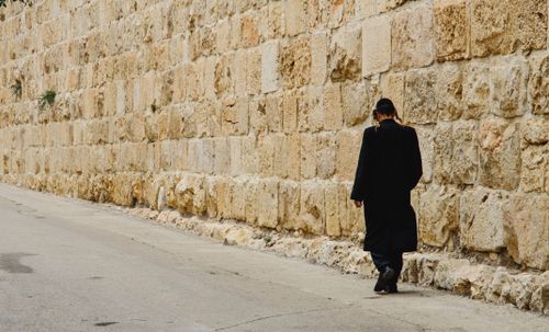 Free Man in black attire walking beside a historic stone wall on a quiet street. Stock Photo