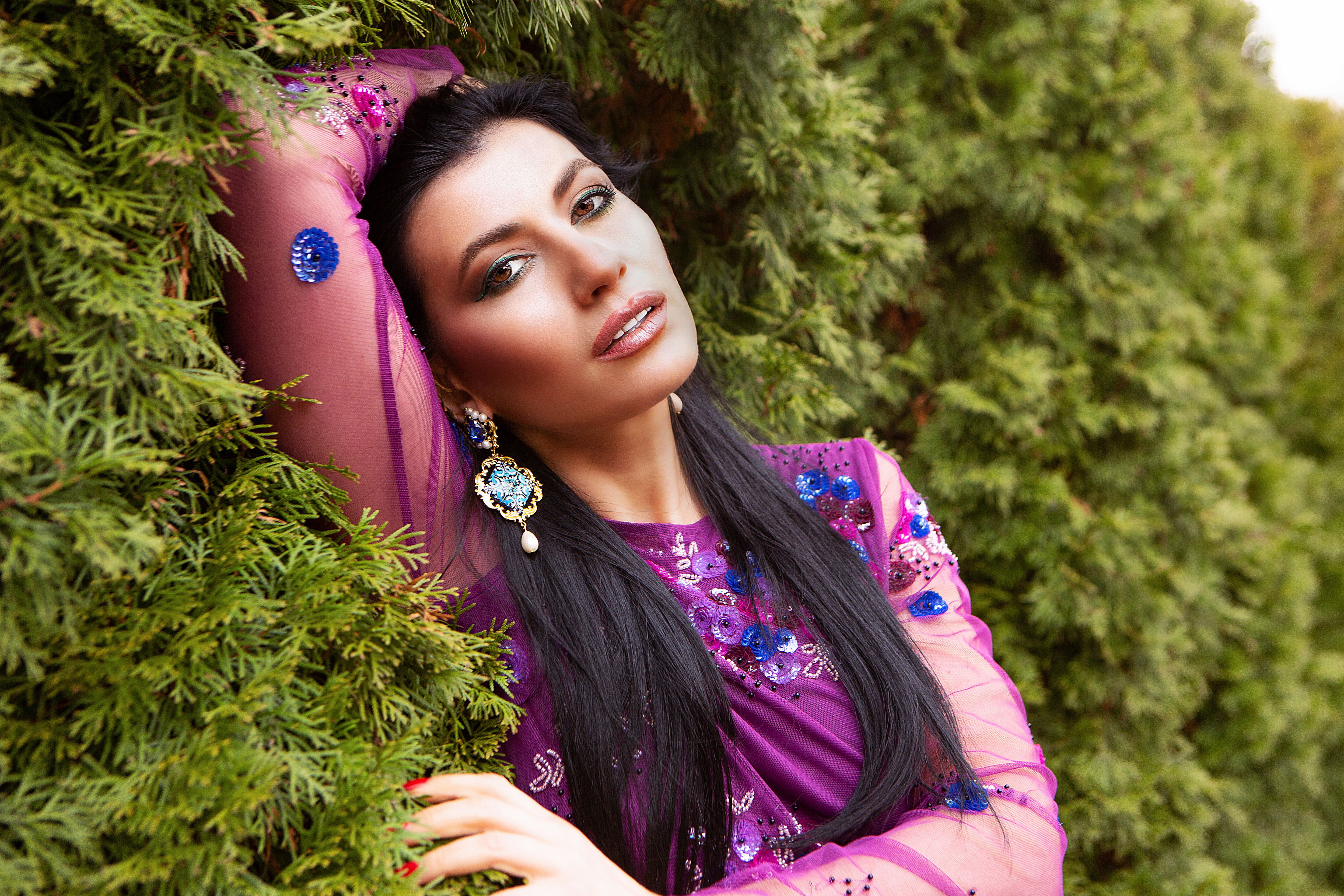 Confident charming female in bright stylish dress with earrings and bright makeup standing near green shrub fence on summer day
