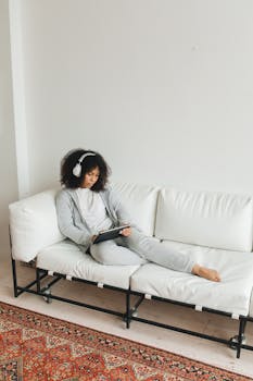 Black woman lounging on a white sofa wearing headphones, using a tablet indoors.