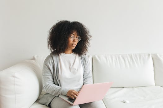 African American woman with glasses using a laptop on a white sofa. Modern lifestyle indoors.