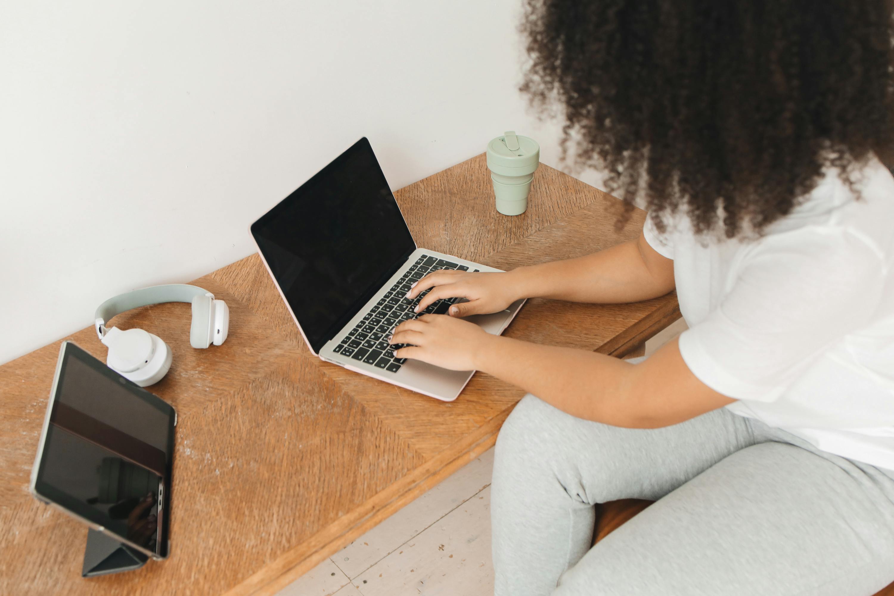Afro-haired woman working remotely on laptop at modern wooden desk with tablet and headphones.