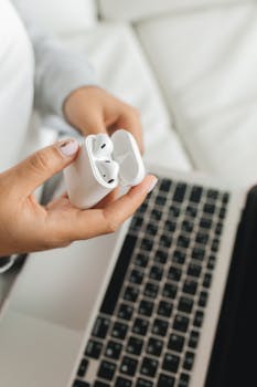 Close-up of a woman holding wireless earbuds above a laptop, depicting remote work or study scenario.