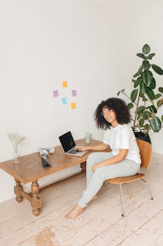 Woman working remotely at home, sitting on a chair with a laptop on a wooden desk.