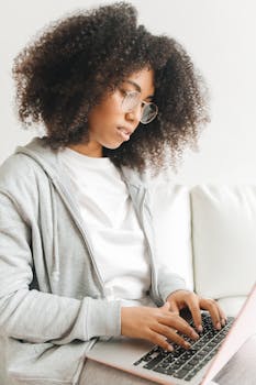Young woman with curly hair typing on laptop at home, focused on remote work.