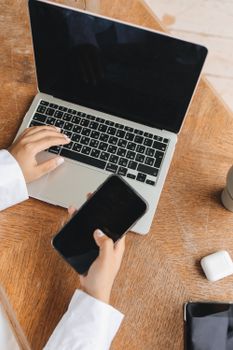 Person using a laptop and smartphone simultaneously at a wooden table.