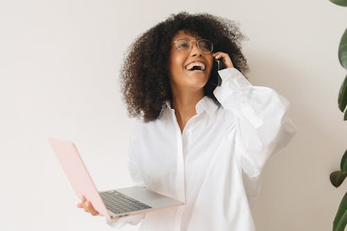 Woman in White Dress Shirt Having a Phone Call while Holding a Laptop