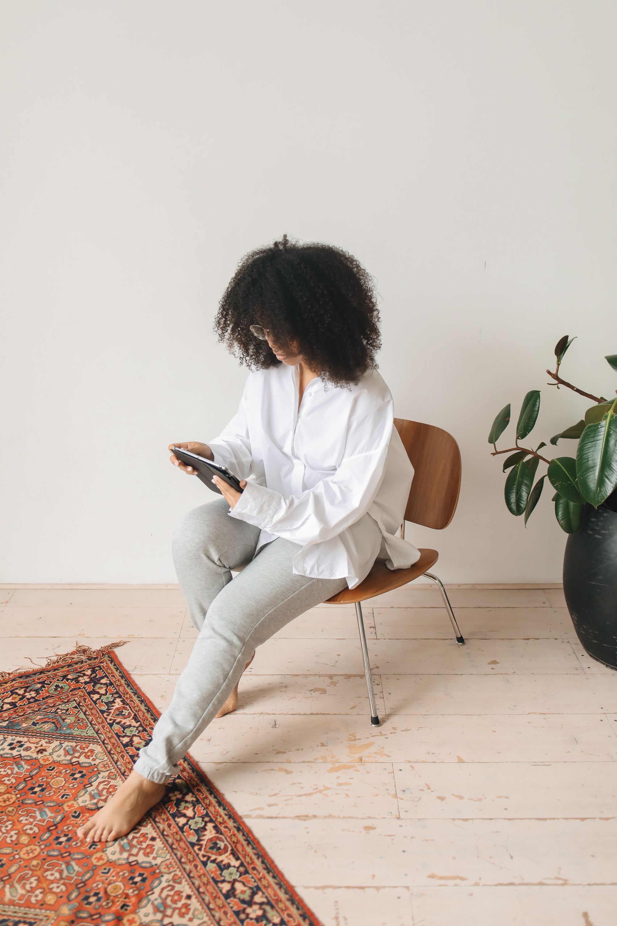 Woman with afro hair sitting on a chic chair, browsing a tablet in a minimalist room.