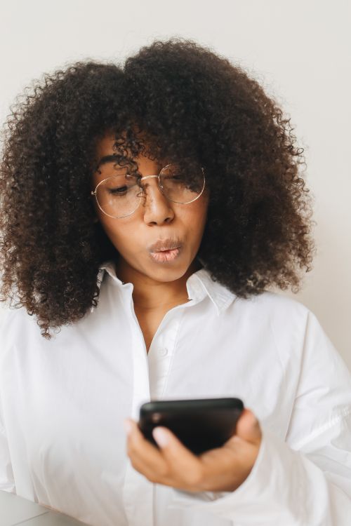 Free A young woman with afro hair and eyeglasses looking at her smartphone indoors. Stock Photo