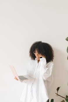 Confident woman multitasking with laptop and phone in bright, minimalist office space.