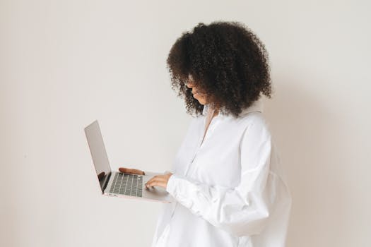 African American woman in white shirt working remotely with laptop, side view.