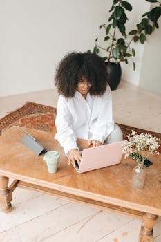 Woman working from home with pink laptop, tablet, and indoor plants on a cozy desk.