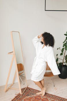 A woman in a white bathrobe poses confidently in front of a full-length mirror indoors.