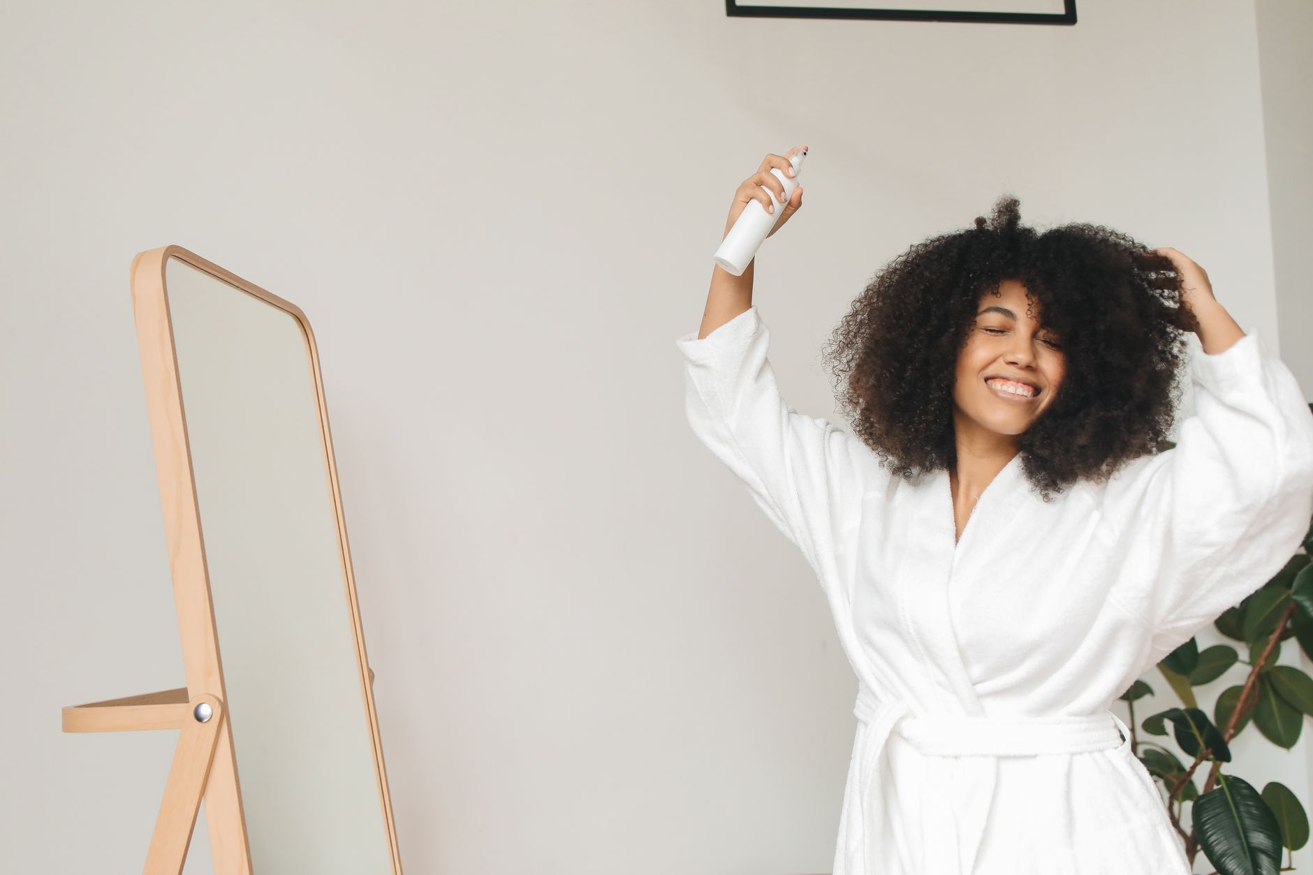 Smiling woman in a bathrobe applying hair spray at home, enjoying self-care routine indoors.