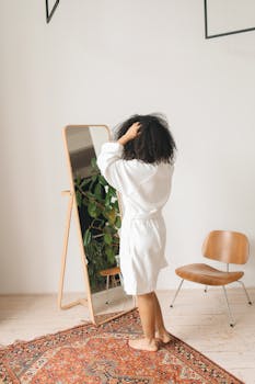 Woman in white bathrobe adjusting her curly hair in front of a mirror in a cozy home setting.