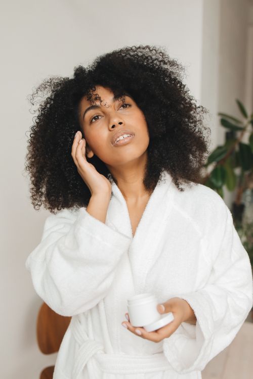 Free A woman wearing a bathrobe applies facial cream indoors with natural lighting. Stock Photo