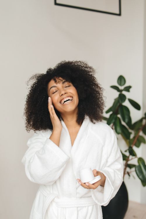 Free A joyful woman in a white bathrobe applying face cream indoors. Stock Photo